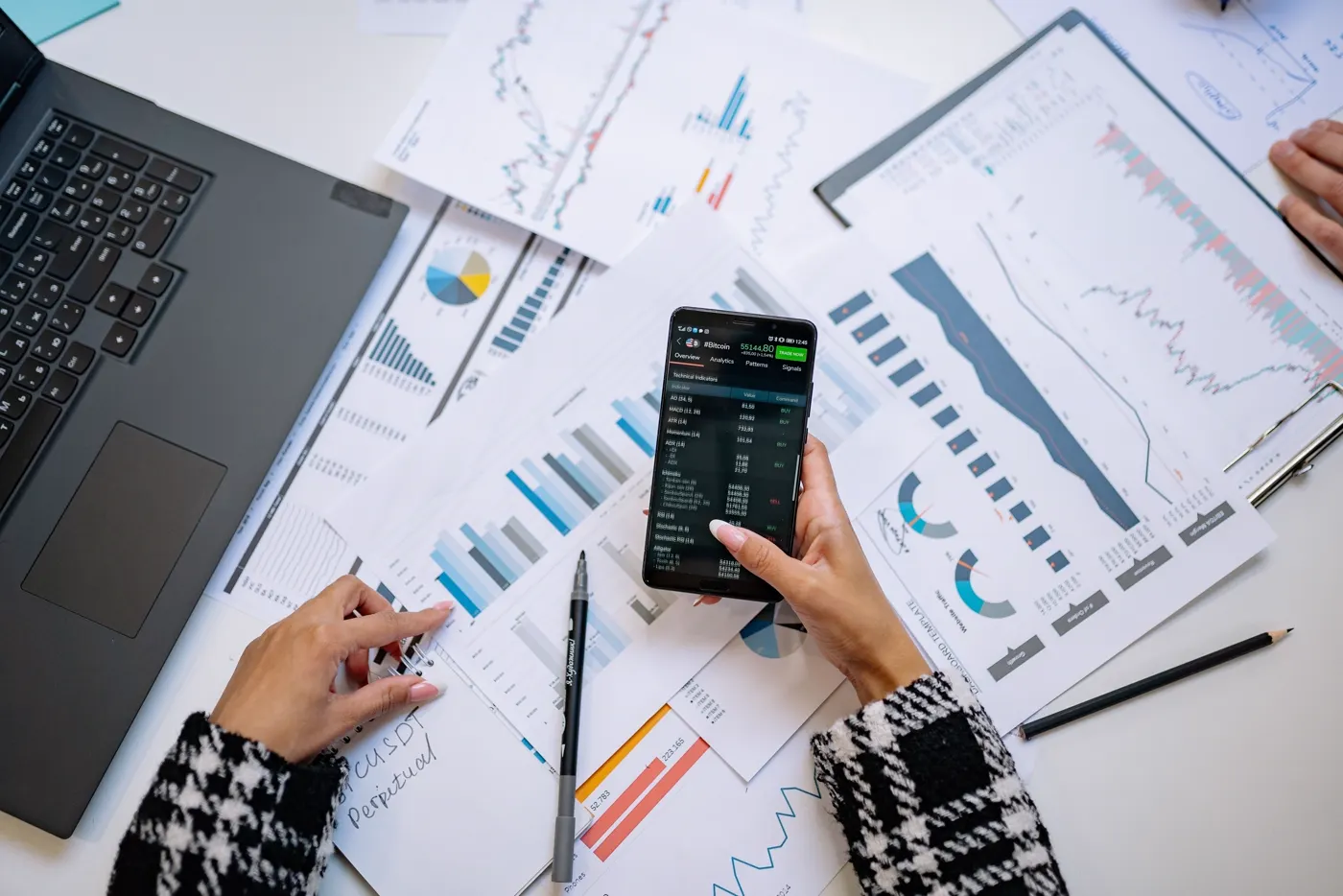 Escritorio cubierto de informes impresos con gráficos y tablas mientras una persona revisa datos en su teléfono